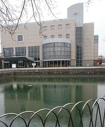 手前にフェンスなどで囲まれた大きな池があり、後方にクリーム色やグレー色のさざんかホール外観が建っている写真