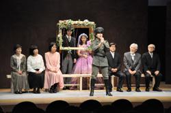 植物や花であしらわれた額の中に、タキシードを着た男性とピンク色のドレスを着た女性が顔だけ出し、前に並べた長椅子の左側に3名の女性、右側に3名の男性が座り、前中央に軍服を着た男性がマイクを持って話している一場面の写真