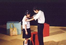 赤色や水色の椅子が置いてある前で、左側の制服姿の女性がうつむき、右側の男性が女性の両肩に手を置いている一場面の写真