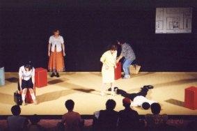 赤色の椅子に制服姿の女性が座り、3名の女性が登場し黄色のシャツを着た女性の足元に、制服姿の男性がうつ伏せで足を広げて倒れている一場面の写真