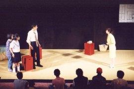 左側の赤色の椅子を挟んで、制服姿の男女と女性が立ち、右側の黄色のシャツを着た女性と向かい合って話しをしている一場面の写真