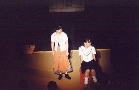 右側の赤色の椅子に腰かけた制服姿の女性と、中央に立つ女性が演劇を行っている一場面の写真
