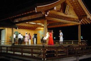 能舞台の上で、7名の人達が後ろを向いているなか、赤色のドレスを着た人と白色のエプロンを着用した人が向かい合っている一場面の写真