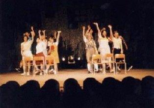 学校の椅子が2か所にまとまって設置された場所に6名ずつ立ち、右手を上げている一場面の写真