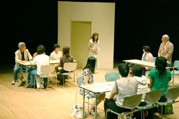 長机の横に立つ白髪の男性と中央に立つ女性が向かい合っているのを、長机をつけて置いた椅子に座っている人達が見ている一場面の写真