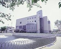 手前に噴水があり左側に入り口、右側に円形の全面ガラス張りの窓のある4階建てのさざんかホールの建物外観の写真