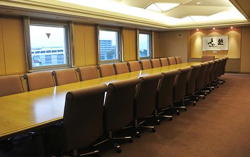 奥に3つの四角形の窓があり、長いテーブルの両脇に椅子が設置されている会議室の写真