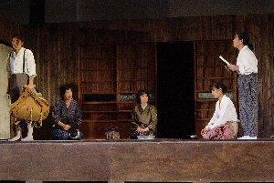 昔の民家の中で、もんぺ姿の3名の女性が間隔をあけて正座し、右側の女性が立って、左側のリュックを持って立っている男性に向かって話している一場面の写真