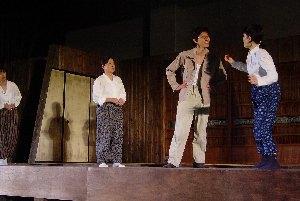昔の民家の中で、右側のもんぺ姿の女性と男性が向かい合って立ち話しをしているのを、左側に立つ2名の女性が見ている一場面の写真