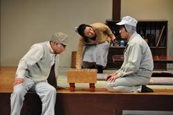碁盤の右側に作業着を着た男性が正座で座り、左側にベストを着た男性が碁盤を見て、中央の女性が正座をしている男性の顔を覗いている一場面の写真