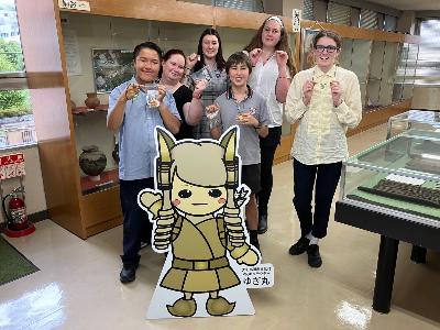 リズモー学生が文化財のマスコットキャラクターゆぎ丸と一緒に作成した勾玉を掲げながら記念撮影している様子
