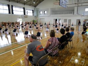 椅子に座ったリズモー市の学生や関係者が、体育館で床に寝て両足を揃えて持ち上げている生徒たちの演技を見ている写真