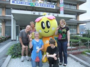 リズモー市の学生3人と関係者の男性と女性が、市役所の前でマスコットキャラクターのみくちゃんと一緒に記念撮影をしている写真