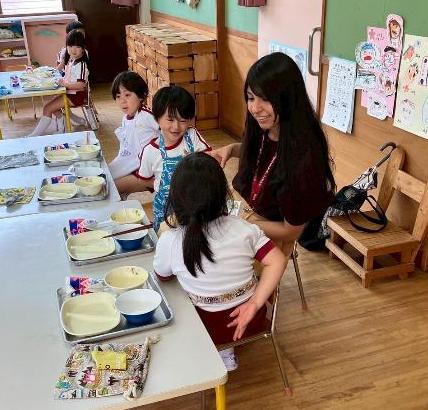 テーブルに座っている子供たちと1人の女性が遊んでいる様子。テーブルの上には給食のお皿やトレイなどが置かれている。