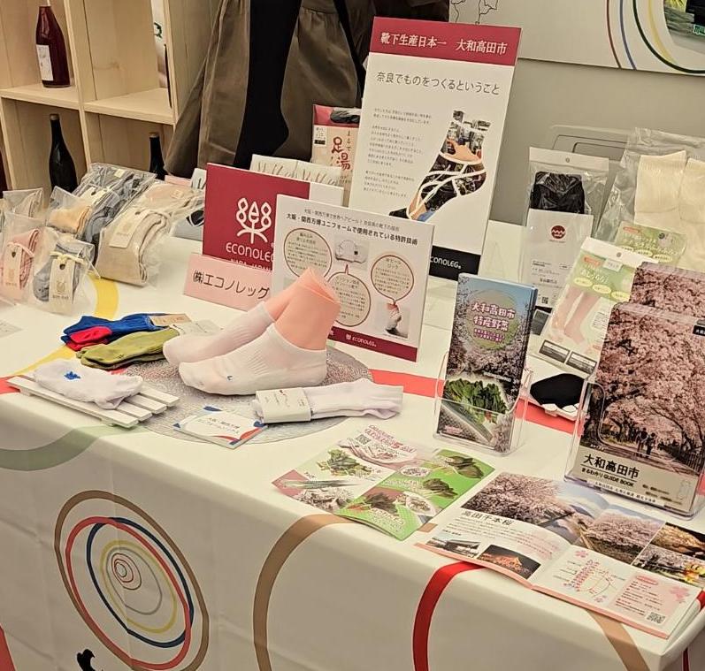 展示のテーブルに靴下の商品、会社のネームプレート、大和高田市のパンフレットが載せている様子。