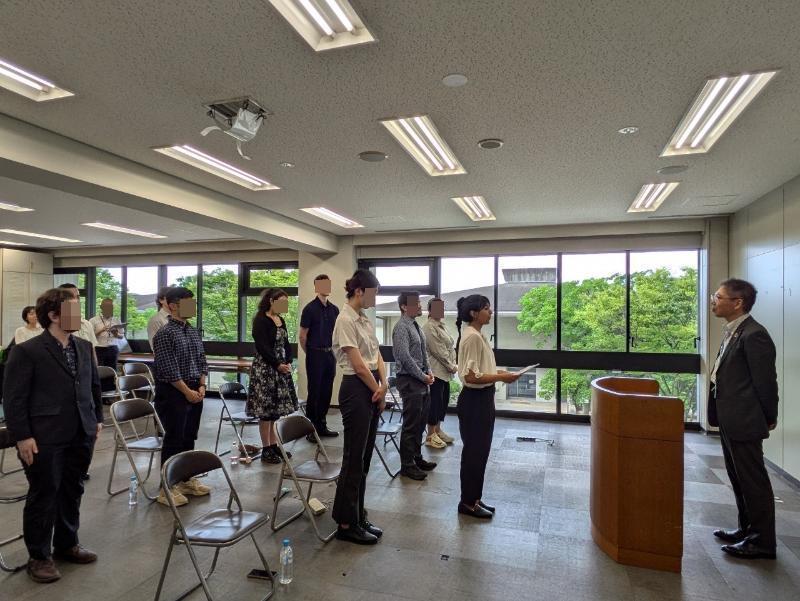 部屋の一番前に立っている男性のほうを向き、椅子の前に立っている9人の男女の様子