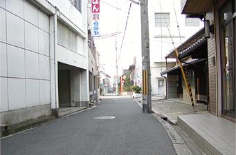 細い道路と敷地の境界線沿いに電信柱が立ち、両脇に建物が並んでいる道路の写真