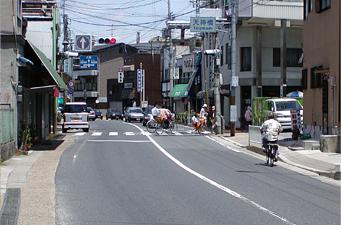 奥に赤信号が見え横断歩道を渡る人達や自転車や車で往来している人達を写した写真