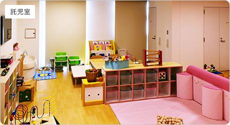 左壁側や中央に棚が設置され、奥に木製のテーブルや低い椅子、手前に遊ぶスペースがある託児室の写真