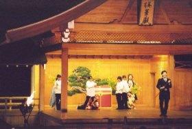 大きな赤色の箱の周囲に5名の人達が立ち、右側に立つ黒色のスーツ姿で杖を持った男性を見ている一場面の写真