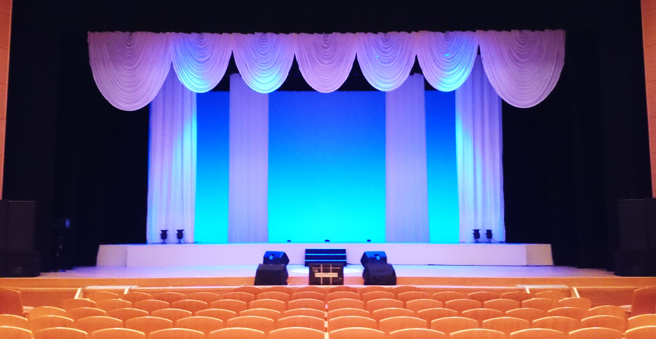 ステージの上に波型に幕が吊るされて青紫色の照明が照らされ、中央左右に小さなスピーカーが設置されている大ホールの写真