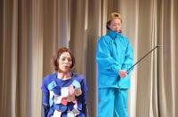 ステージの幕の前で、青色の服を着た女性が座り、右側に水色のジャージ姿の女性棒の様な物を両手で持っている一場面の写真