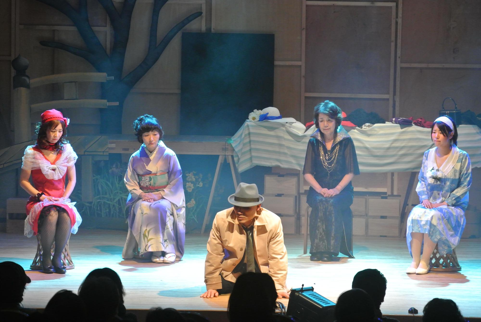 4名の派手な衣装を着た女性が横一列に椅子に座っている中央で、帽子を被ってベージュ色のコートを着ている男性が座って床に手をついているのを見ている一場面の写真