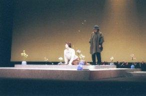 ステージ上の四方に花が生けられた花瓶が置いてある四角形の土台の上の中央に、両手を床について座っている女性と、右側の帽子を被った男性が見ている一場面の写真