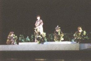 ステージ上に設置された四角形の土台の上に、植物が絡まった4名の人達が間隔をあけて座り、中央に女性が立っている一場面の写真