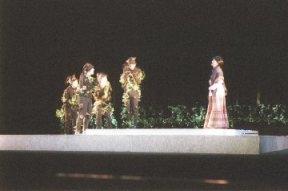 ステージ上に設置された四角形の土台の上に、左側の植物が絡まった4名の人達と右側の女性と向かい合って立っている一場面の写真