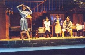 洋館の建物内を舞台に、左手前の水色のワンピースを着た女性が立って頭に被っている帽子に両手を添えているのを、椅子に座っている男女と間に立っている女性が見ている一場面の写真