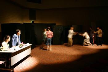 ステージの左側の長机に女性と中腰に立った男性がおり、中央の2名、右側の4名の人達が演劇を行っている一場面の写真