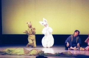 舞台の上で、狸と狐の衣装を着た人が床に膝をついて両手を合わせている一場面の写真