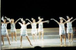 ステージの上で、白色の短いドレスを着た女性達が両手を広げて踊っている一場面の写真