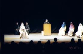 中央の壇上に黒色のマントを羽織った人が立ち、左側に黒色のマントを羽織った3名の人達や右側の頭をショールで覆った3名の女性が椅子に座り、左側に全身白色の服で覆われた人が座っている一場面の写真