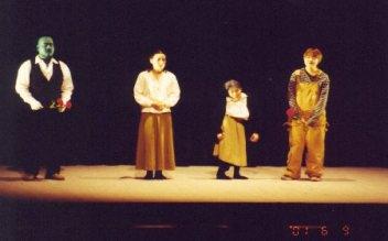 ステージ上に左からフランケンシュタインに模した男性、両手を握りしめている女性、小さな女の子、オーバーオールを着た人が横一列に並んで演技をしている一場面の写真