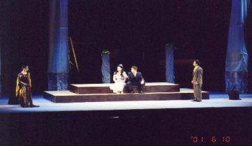 ステージ上に設置された2段のひな壇に女性と男性が座り、両端に2名の男性がそれぞれ立っている演技を行っている一場面の写真
