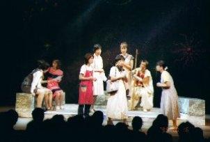 ステージ上に設置された半円形の石材の土台上の中央に、右手に杖を持った白装束の人が石材に座り、8名の女性が回りに立ったり座ったりして演劇を行っている一場面の写真