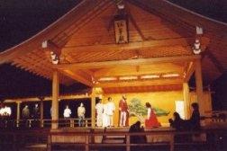 能舞台の上で、赤色のマントを羽織った男性の右側で向かい合って膝まづいている人が演技を行っている一場面の写真