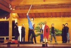 赤色のマントを羽織った人と他6名の前で、青色の服を着た男性が右手で剣を持ち上げている一場面の写真