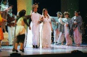王冠を頭に被った男性と白色のドレスを着た女性が腕を組んで立ち、周りの人達が祝福している一場面の写真