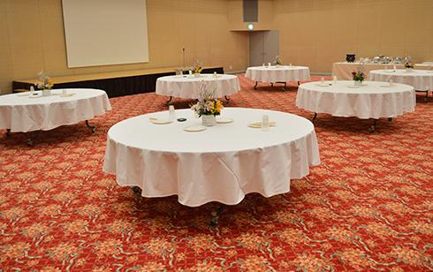 左奥に小さなステージがあり、手前に6つの白色の布がかけられている円形のテーブルが設置されているホール内の写真