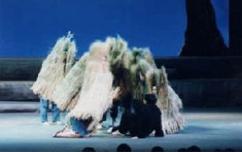 ステージ上に、学生服を着た男性が観客側を背にして膝を立てて座り、藁を背負った人達が男性を囲むように立っている演劇の一場面の写真