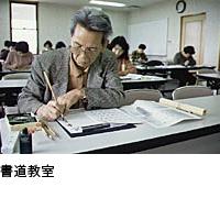 白髪の男性が右手に筆を持ち、半紙に左手を添えて書いている写真