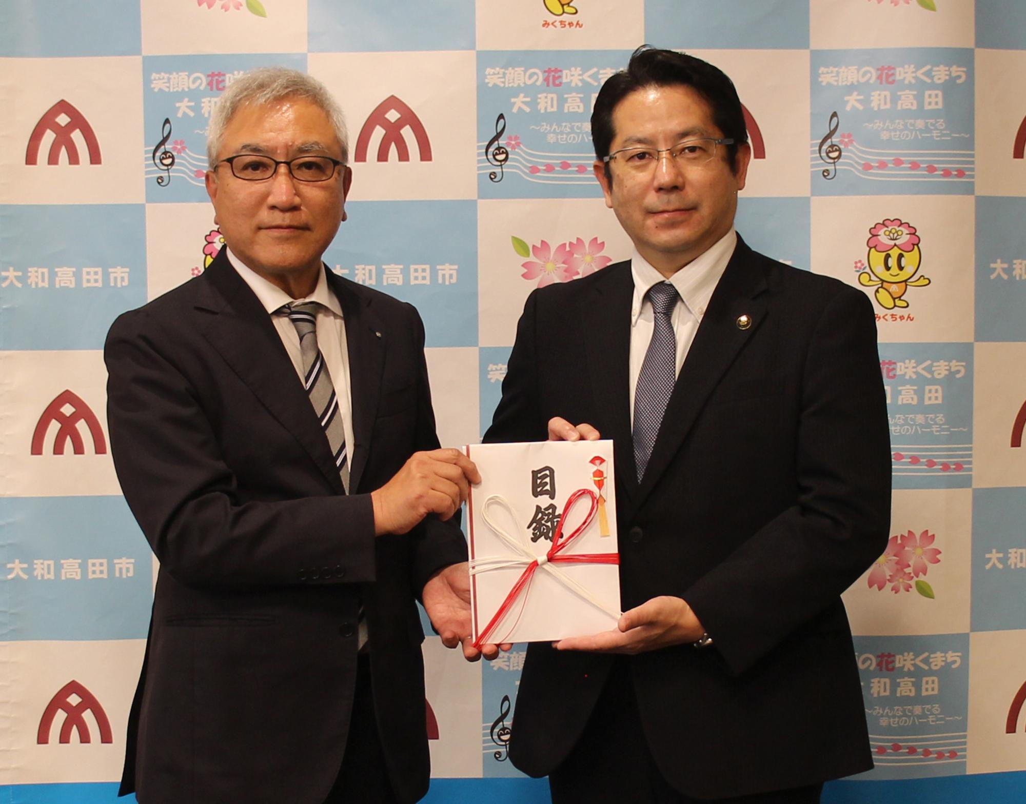 画面左側の奈良ヤクルト販売株式会社谷川代表取締役社長から画面右側の堀内大和高田市長へ寄附金目録を贈呈する。