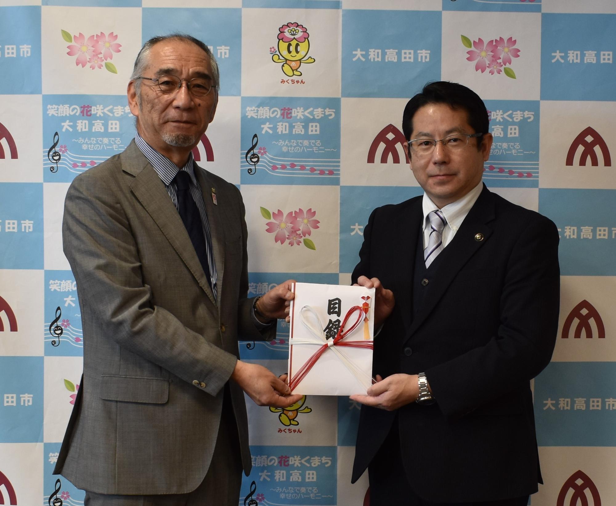 画面左側の高田鋼材工業株式会社の松村代表取締役社長から画面右側の堀内大和高田市長へ寄附金目録を贈呈する。