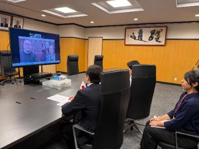 市長、副市長、職員がテレビ画面に向かって椅子に座り、オンライン会議を行っている様子。画面にはリズモー市長と女性が表示されている。