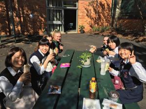 庭に置かれた緑の木造テーブルでパンを食べている女子学生と外国人の女子学生の写真