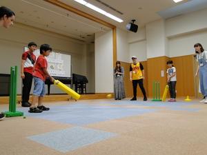 子ども2人が黄色のバットのようなものを持ちボールを打ち合っているのを派遣学生5名が見ている写真
