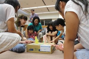 派遣学生と子供達が箱に置いた紙相撲で遊んでいる写真